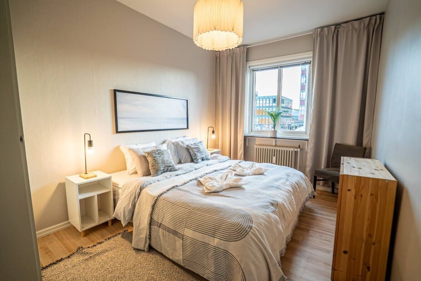 A modern bedroom with a bed, bedside tables, a window with curtains, a radiator, and a view of the city.