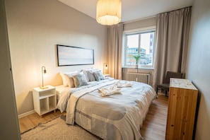 Un dormitorio moderno con cama, mesitas de noche, ventana con cortinas, radiador y vistas a la ciudad.