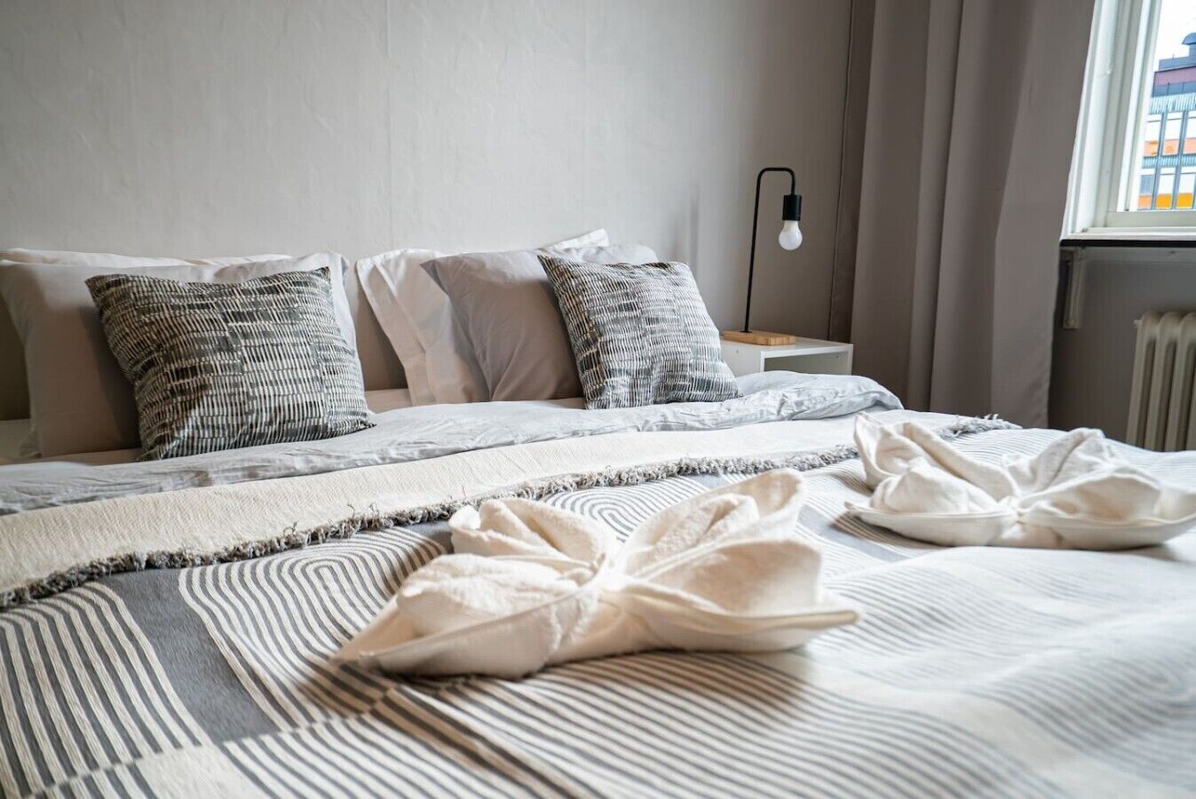 A neatly made bed with striped bedding and multiple pillows, a bedside lamp, and a view of a building through a window.
