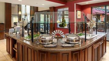 A buffet counter with multiple serving stations, a glass partition, and a view of the hotel lobby.
