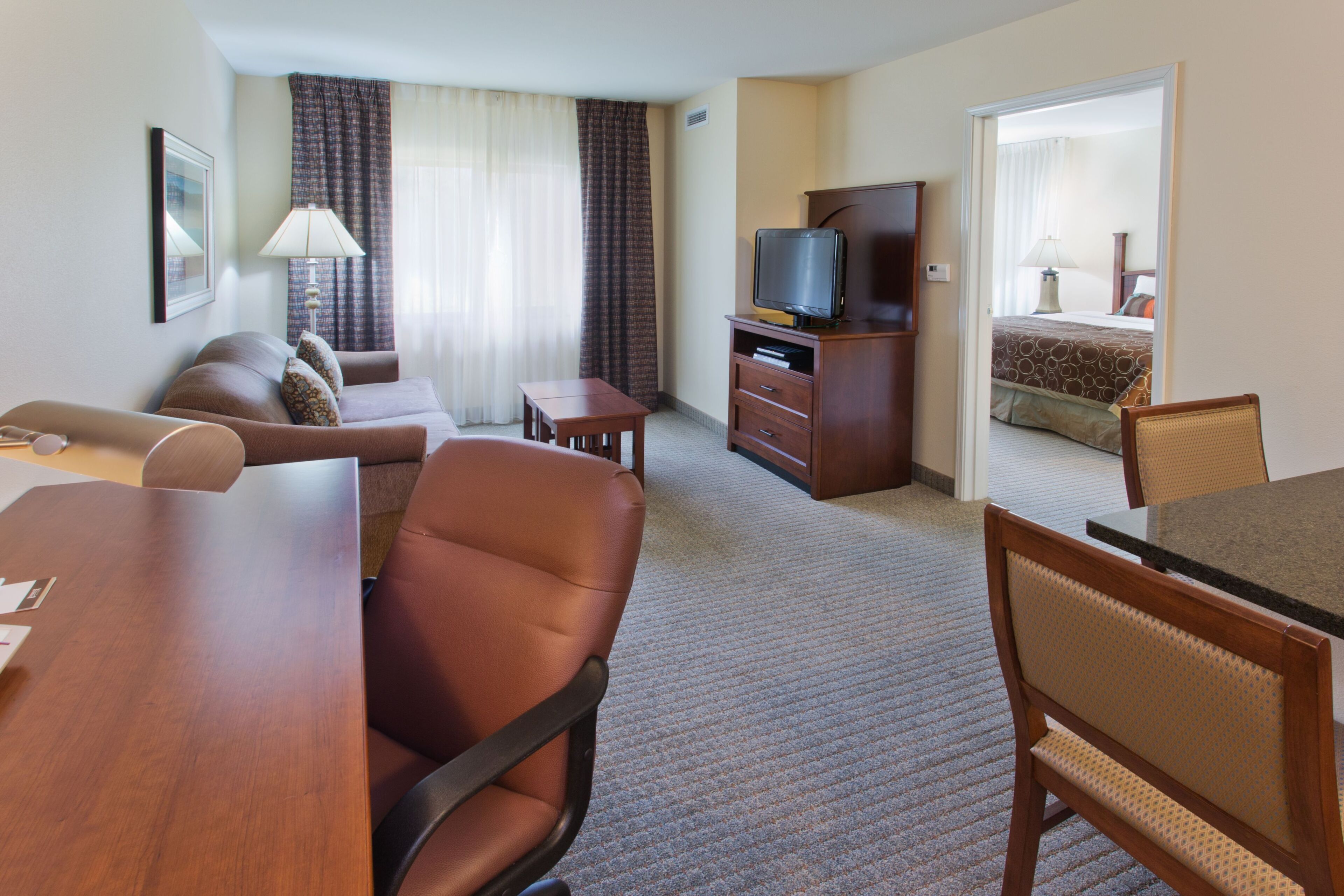 A hotel room with a sofa, a dining table, a TV on a wooden cabinet, and a bed with a patterned bedspread.