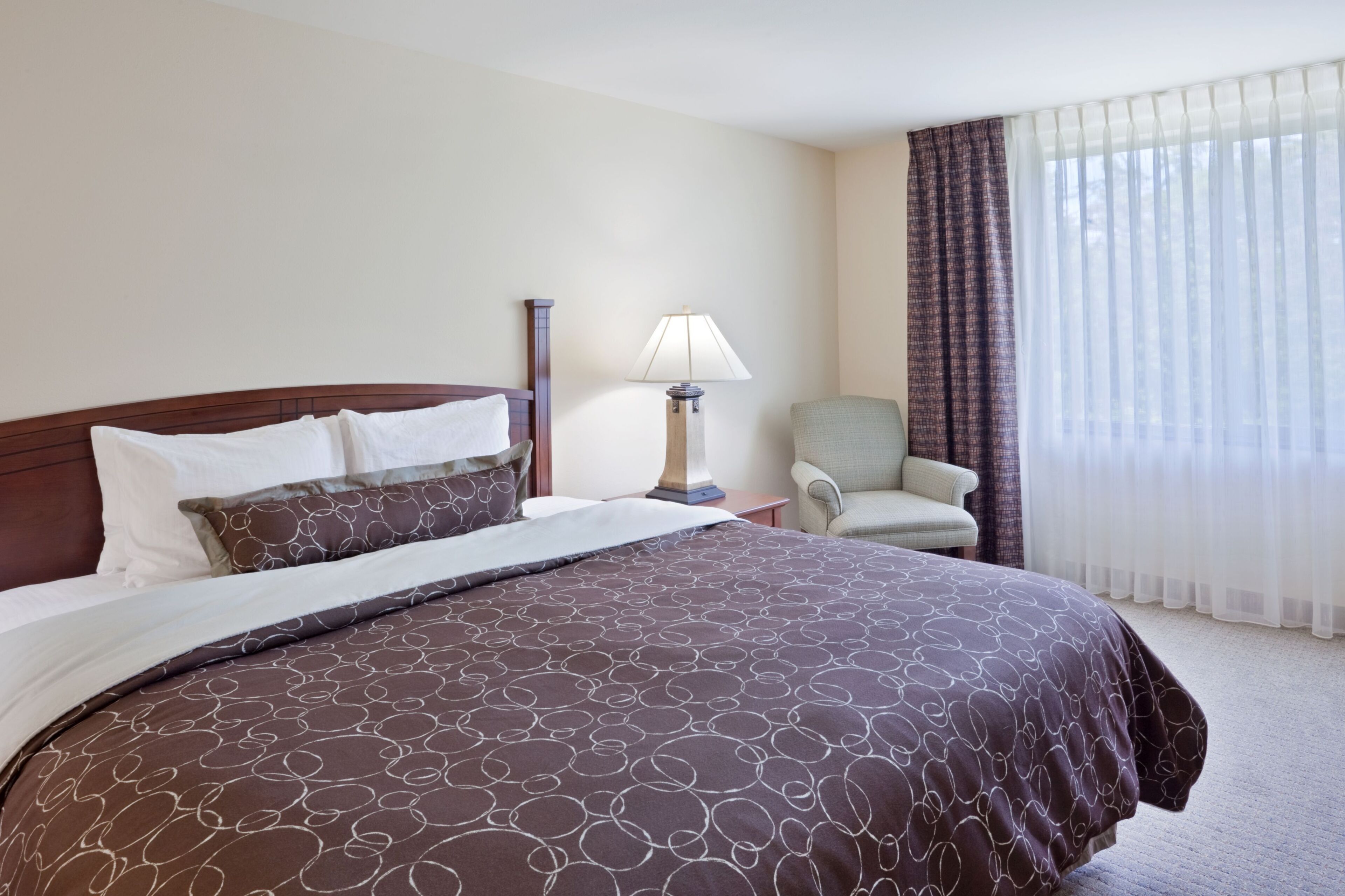A hotel room with a large bed, a bedside table with a lamp, a chair, and a window with curtains.