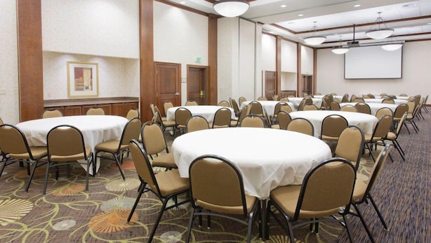 A conference room with multiple round tables and chairs, carpeted flooring, and a projector screen on the wall.