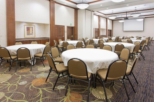 A conference room with multiple round tables and chairs, carpeted flooring, and a projector screen on the wall.