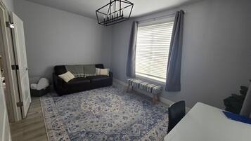 Una habitación con un sofá marrón oscuro, un tapete estampado, un banco, una mesa blanca y una ventana con persianas.