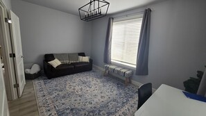 A room with a dark brown sofa, a patterned rug, a bench, a white table, and a window with blinds.