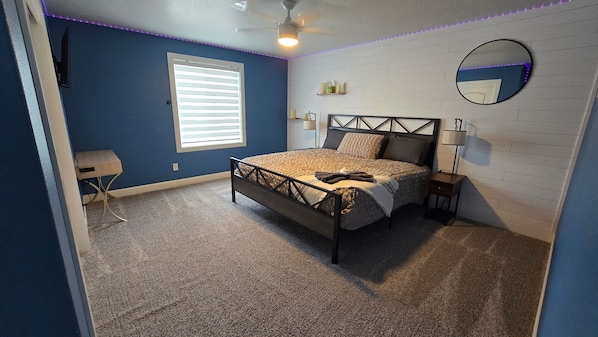 A bedroom with a bed, a ceiling fan, a window with blinds, a bedside table with a lamp, and a wall-mounted mirror.