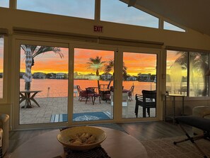Une pièce avec vue sur un coucher de soleil se reflétant sur l’eau, un saladier de fruits posé sur une table et un panneau indiquant la sortie au-dessus de la porte.