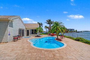 Une piscine à bord incurvé, entourée d’une terrasse dallée. On y trouve des chaises longues et une petite table près de la piscine.