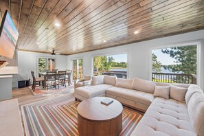 A spacious living area with a large sectional sofa, a dining table with chairs, and a round coffee table. There are large windows providing views of a lake and outdoor deck.