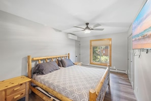 A bedroom with a wooden bed, a ceiling fan, a window with a view, and a wall-mounted air conditioner.