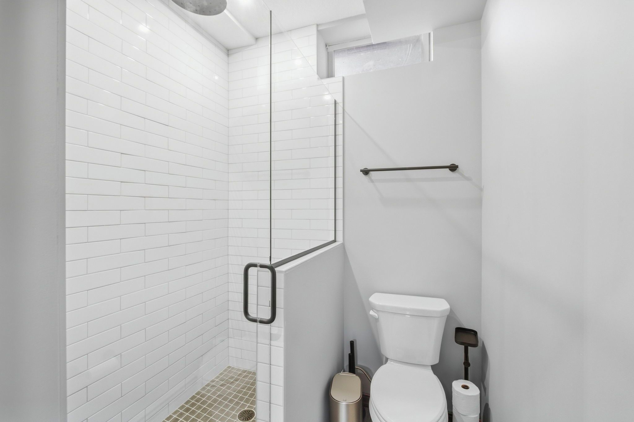 Ein modernes Badezimmer mit weißen Fliesen im U-Bahn-Stil, einer Glasdusche, einer Toilette und einem Mülleimer.