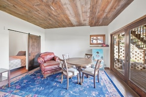 A room with a wooden ceiling, a dining table with chairs, a red leather chair, a bed with patterned bedding, and a window with a view of a brick wall.