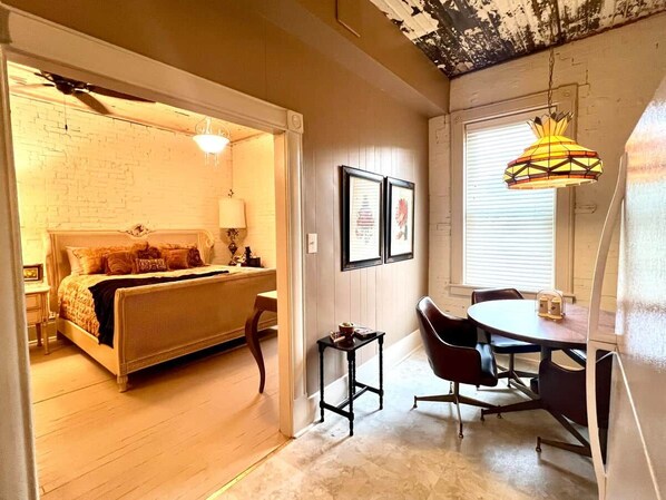 A bedroom with a bed, a bedside table, a chair, a small table, and a window with blinds.