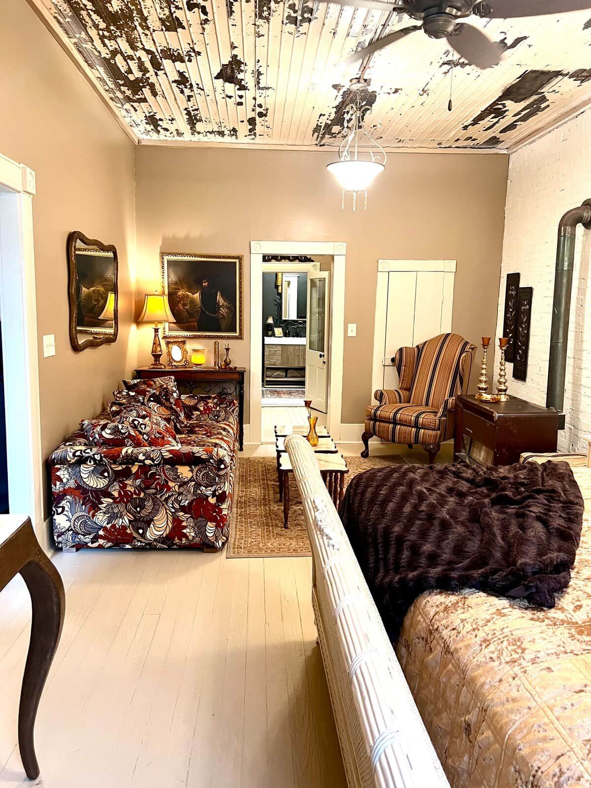 A living room with a sofa, armchair, and a large rug. There is a door leading to a kitchen, and a ceiling fan is visible.