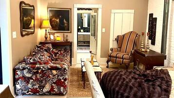 A living room with a sofa, armchair, and a large rug. There is a door leading to a kitchen, and a ceiling fan is visible.