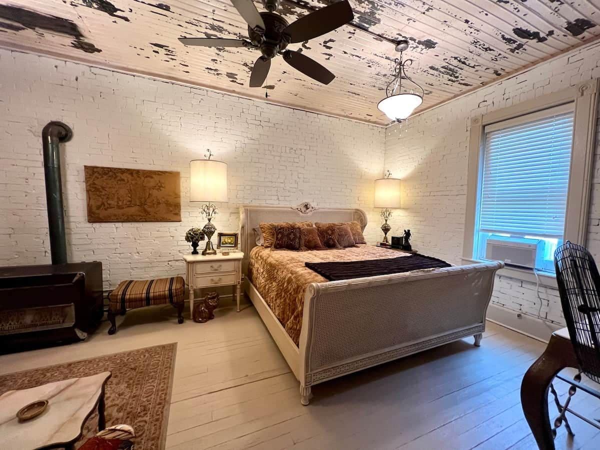 A bedroom with a bed, two bedside tables, a ceiling fan, a window with blinds, and a brick wall.