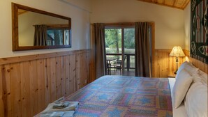 Une chambre avec un mur lambrissé en bois, un grand lit recouvert d’une courtepointe à motifs, une table de chevet avec une lampe, et une vue sur l’extérieur par une fenêtre.