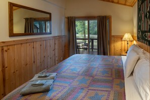 A bedroom with a wooden paneled wall, a large bed with a patterned quilt, a nightstand with a lamp, and a view of the outdoors through a window.