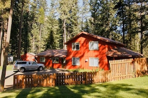 A two-story red wooden cabin with a fenced yard, surrounded by tall trees.