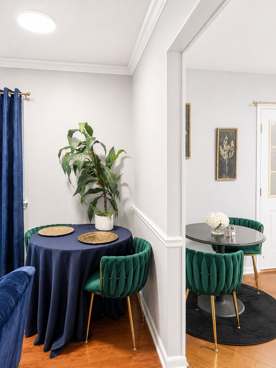 Ein Essbereich mit zwei runden Tischen, jeder mit dunkelgrüner Tischdecke und goldfarbenen Tellern. Zwei grüne, gepolsterte Stühle mit goldenen Beinen stehen davor. Auf dem linken Tisch steht ein Blumentopf, und ein gerahmtes Bild hängt an der Wand.