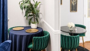 Un comedor con dos mesas redondas, cada una con mantel oscuro y platos con borde dorado. Hay dos sillas tapizadas en verde con patas doradas. Una planta en maceta está sobre la mesa de la izquierda, y un cuadro enmarcado cuelga en la pared.