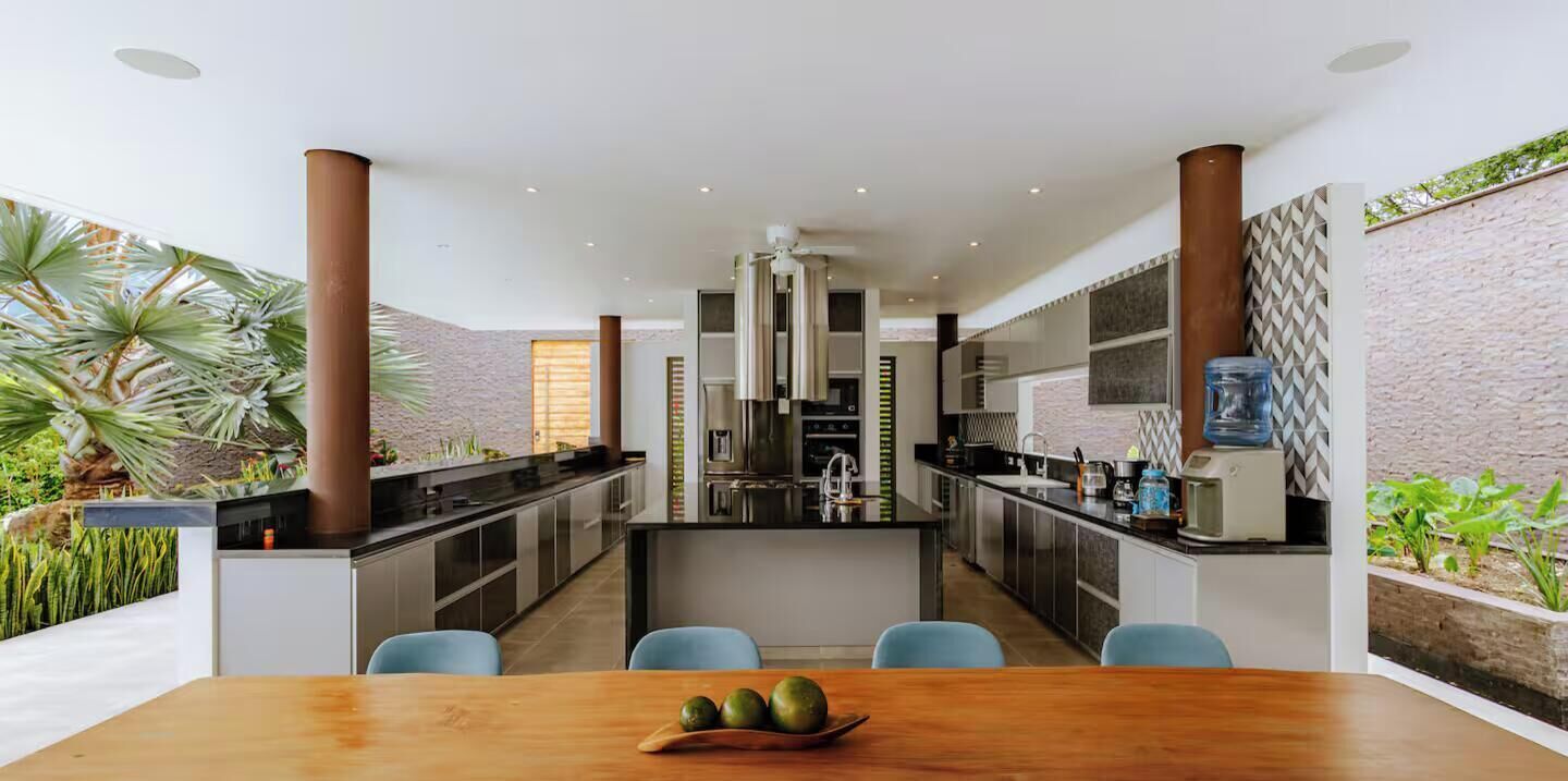 Una cocina moderna con una isla central, encimeras negras y electrodomésticos de acero inoxidable. Hay un comedor con una mesa de madera y sillas azules.