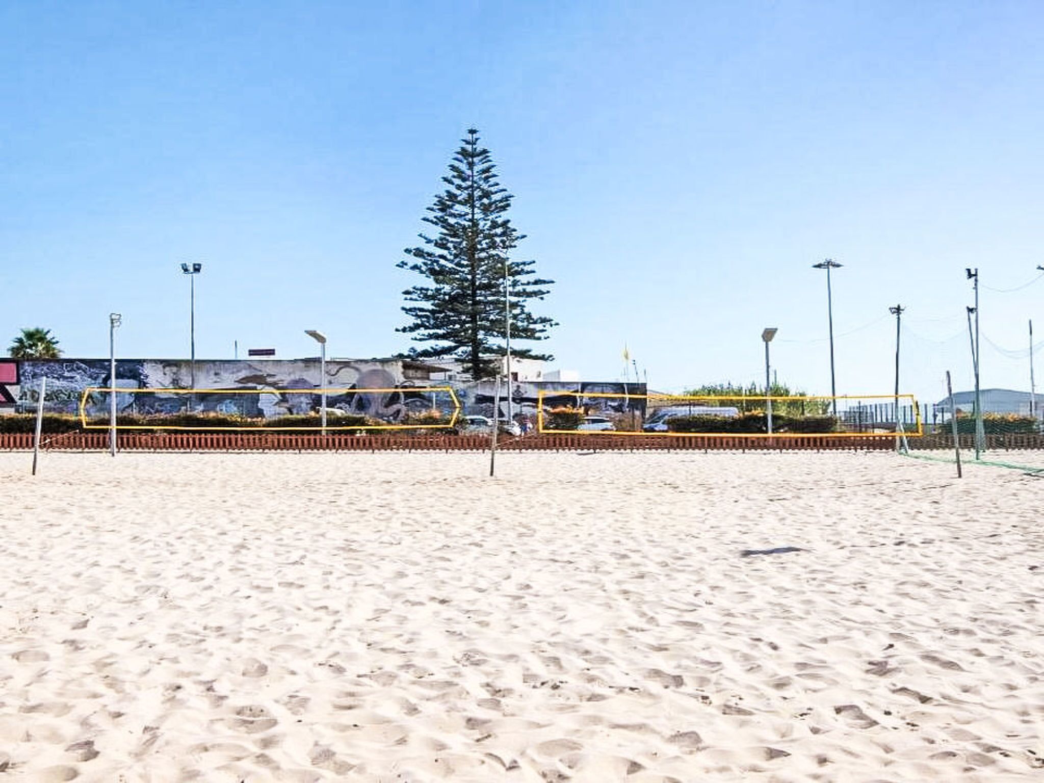 Ein Sandstrand mit einem Volleyballnetz, einem hohen Baum und einem Gebäude im Hintergrund.
