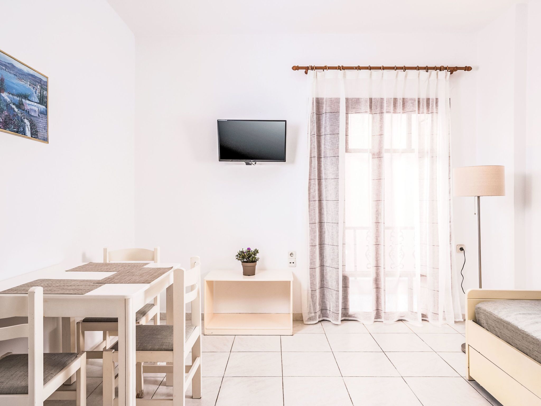 A minimalist room with a flat-screen TV, a dining table with chairs, a bedside table with a lamp, and a window with sheer curtains.