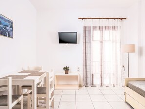 A minimalist room with a flat-screen TV, a dining table with chairs, a bedside table with a lamp, and a window with sheer curtains.