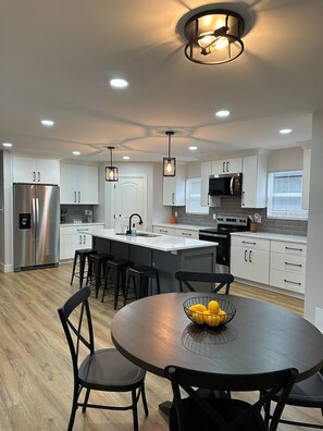 A modern kitchen with white cabinets, a stainless steel refrigerator, a built-in oven, and a dining area with a round table and chairs. - New Modern 4-bedroom 3-Bath Home near downtown Indianapolis and Speedway (Indianapolis)