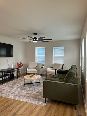 A living room with a ceiling fan, two windows with blinds, a green sofa, a beige armchair, a coffee table, and a TV mounted on the wall. - New Modern 4-bedroom 3-Bath Home near downtown Indianapolis and Speedway (Indianapolis)