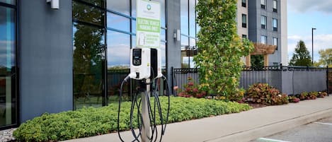 Une borne de recharge pour véhicules électriques, avec un panneau indiquant qu’elle est réservée aux véhicules électriques, se trouve à côté d’un bâtiment moderne doté de grandes fenêtres et d’un arbre.