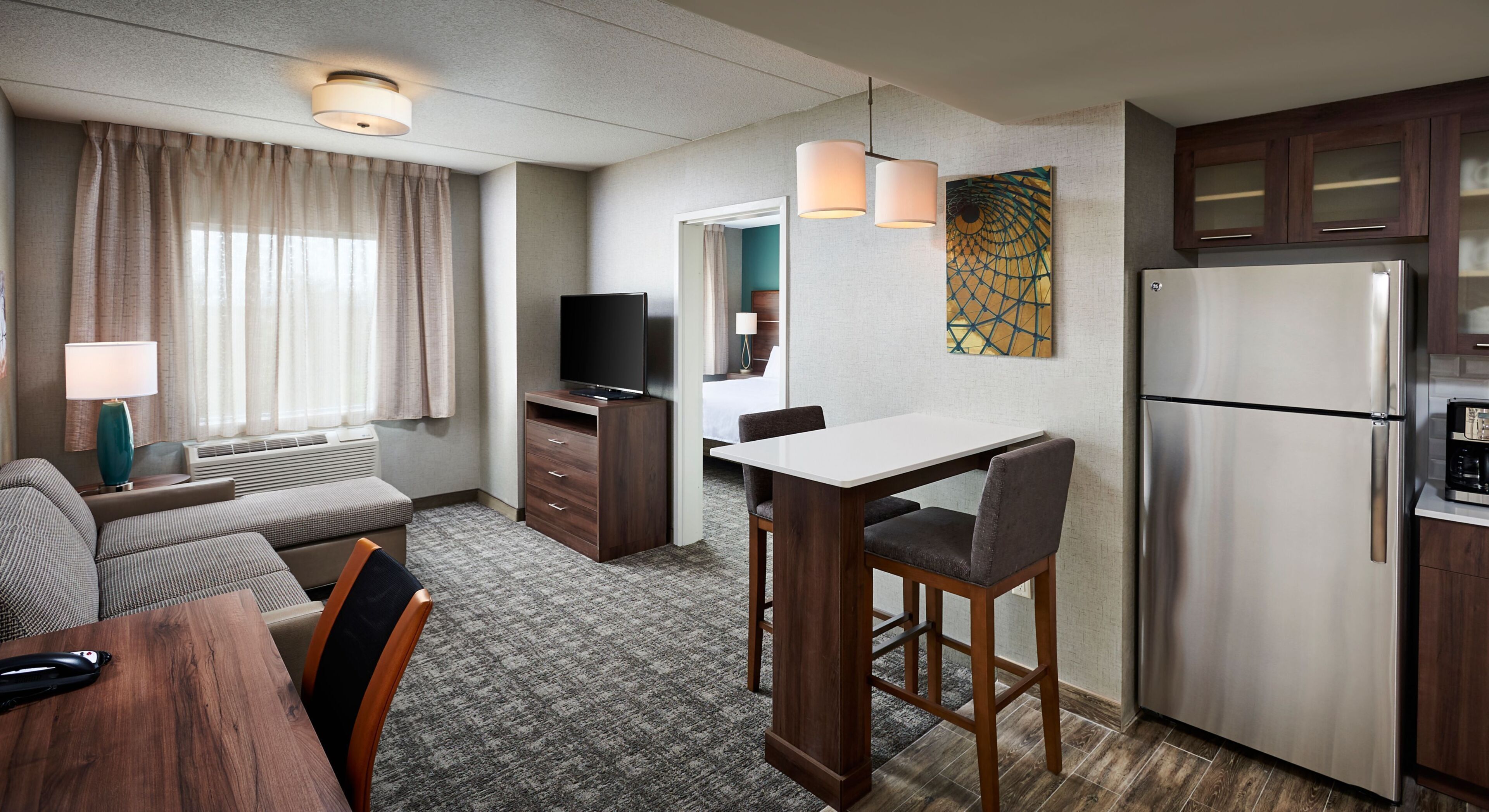 Une chambre d’hôtel équipée d’une kitchenette, d’un coin repas, d’un espace salon avec un canapé et des fauteuils, d’une télévision et d’un lit en arrière-plan.