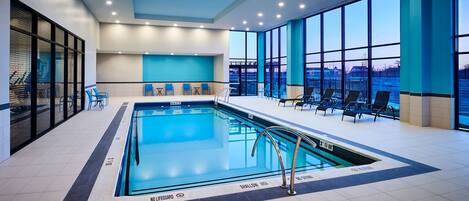 An indoor swimming pool with clear markings, including 'NO LIFEGUARD' and 'SHALLOW AREA' signs, surrounded by lounge chairs and tables.