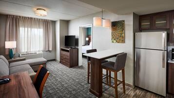 Une chambre d’hôtel équipée d’une kitchenette, d’un coin repas, d’un espace salon avec un canapé et des fauteuils, d’une télévision et d’un lit en arrière-plan.
