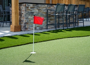 A golf hole with a red flag on a green surface, a stone wall, and outdoor seating.