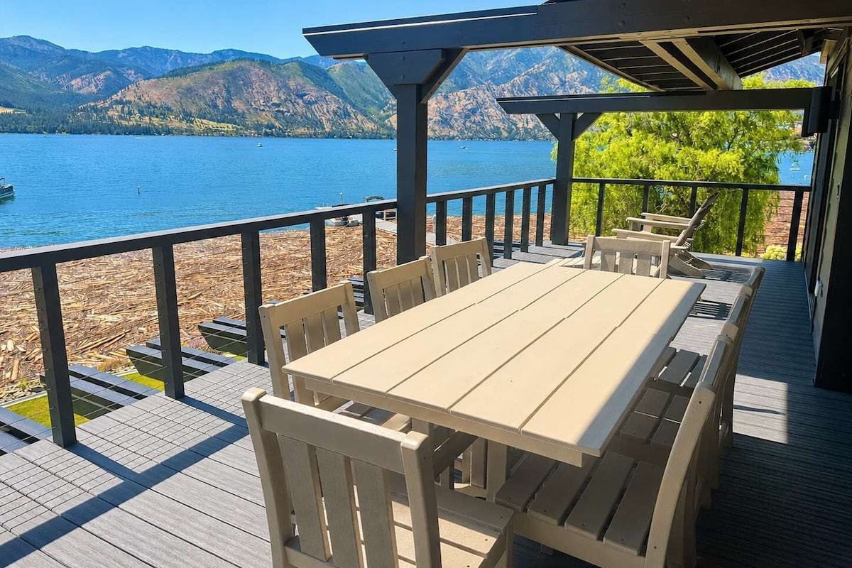 A wooden deck with a table and chairs overlooking a lake with mountains in the background.