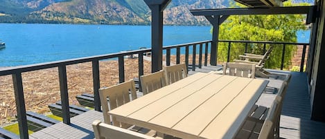 A wooden deck with a table and chairs overlooking a lake with mountains in the background.