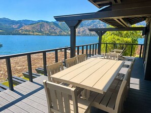 Una terraza de madera con una mesa y sillas, con vista a un lago y montañas al fondo.