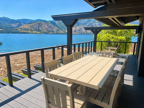 A wooden deck with a table and chairs overlooking a lake with mountains in the background.