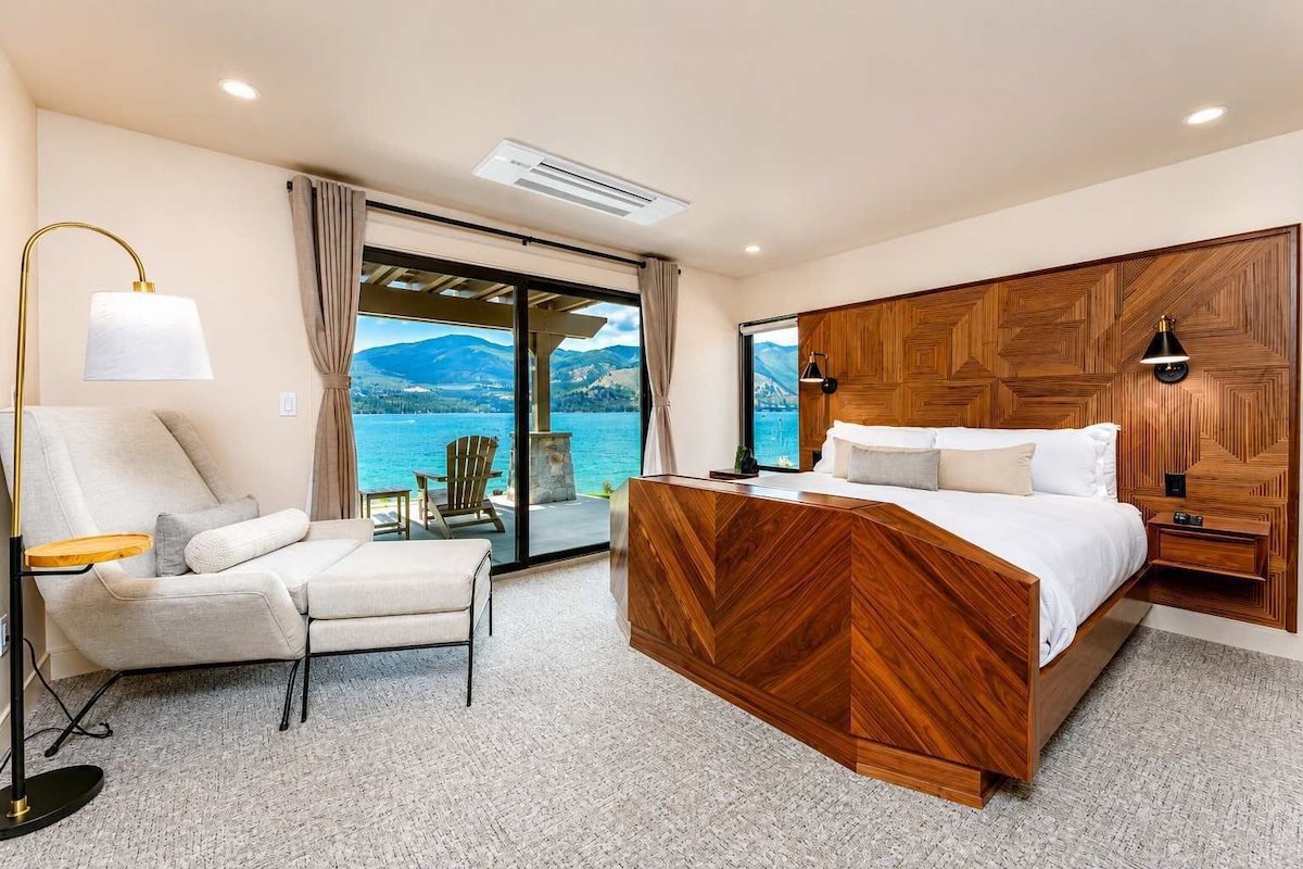 A bedroom with a bed, a chair, a lamp, and a view of a lake.