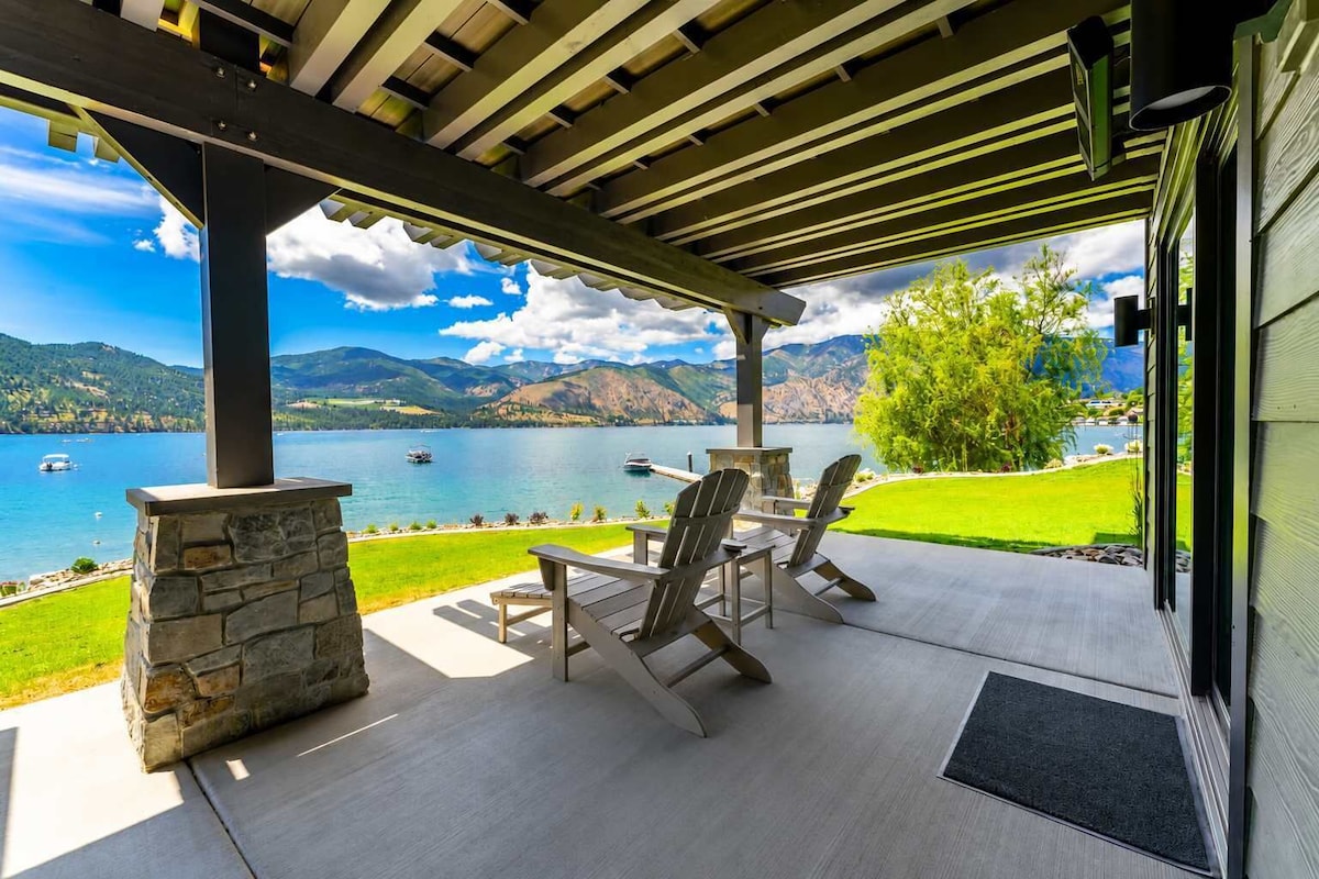 A covered outdoor seating area with a stone fireplace, overlooking a lake with boats and mountains in the background.