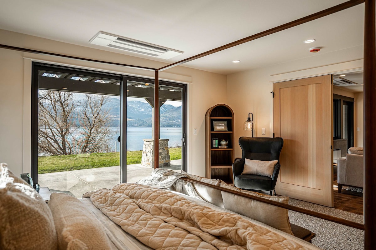 A bedroom with a large bed, a chair, a bookshelf, and a sliding glass door leading to a balcony with a view of a lake and mountains.