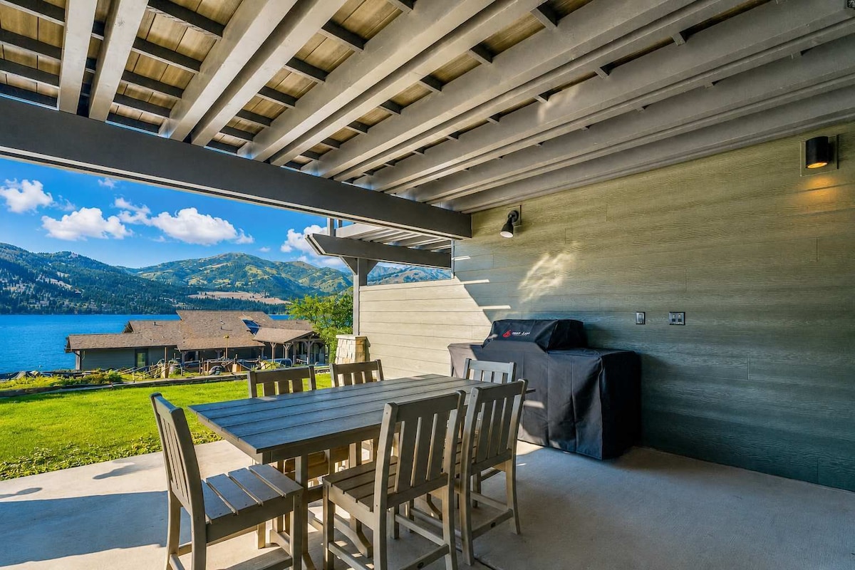 An outdoor patio with a table and chairs, a grill, and a view of a lake and mountains.