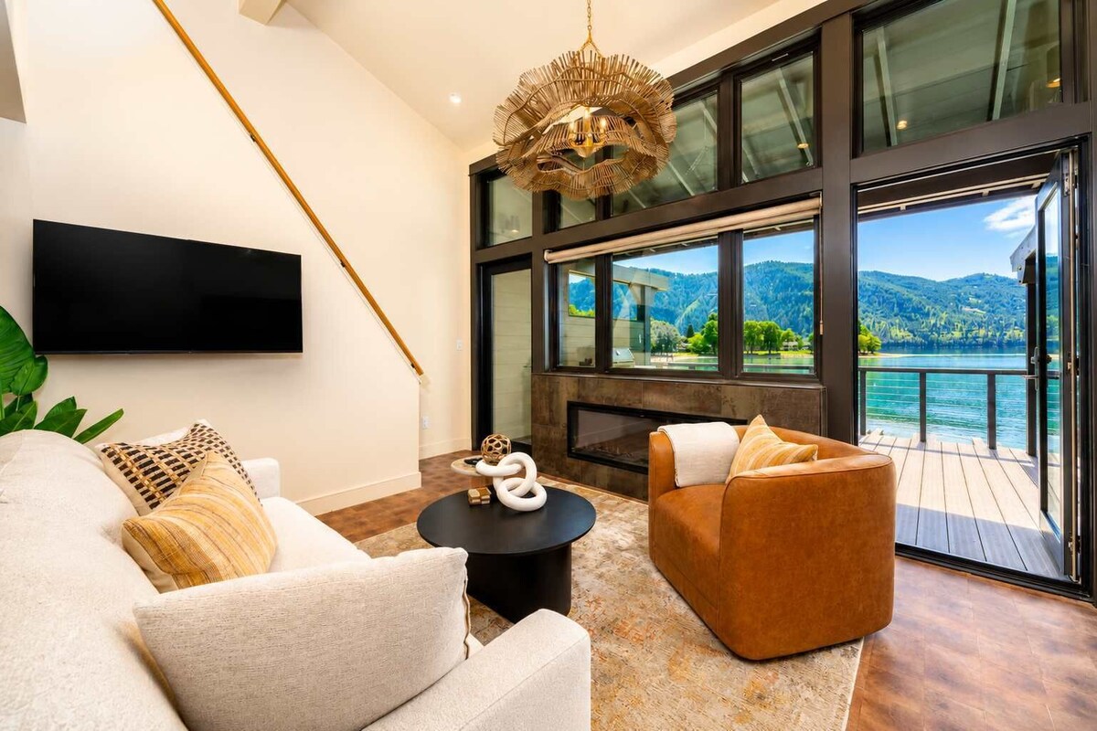 A modern living room with a fireplace, large windows overlooking a lake, and a flat-screen TV mounted on the wall.