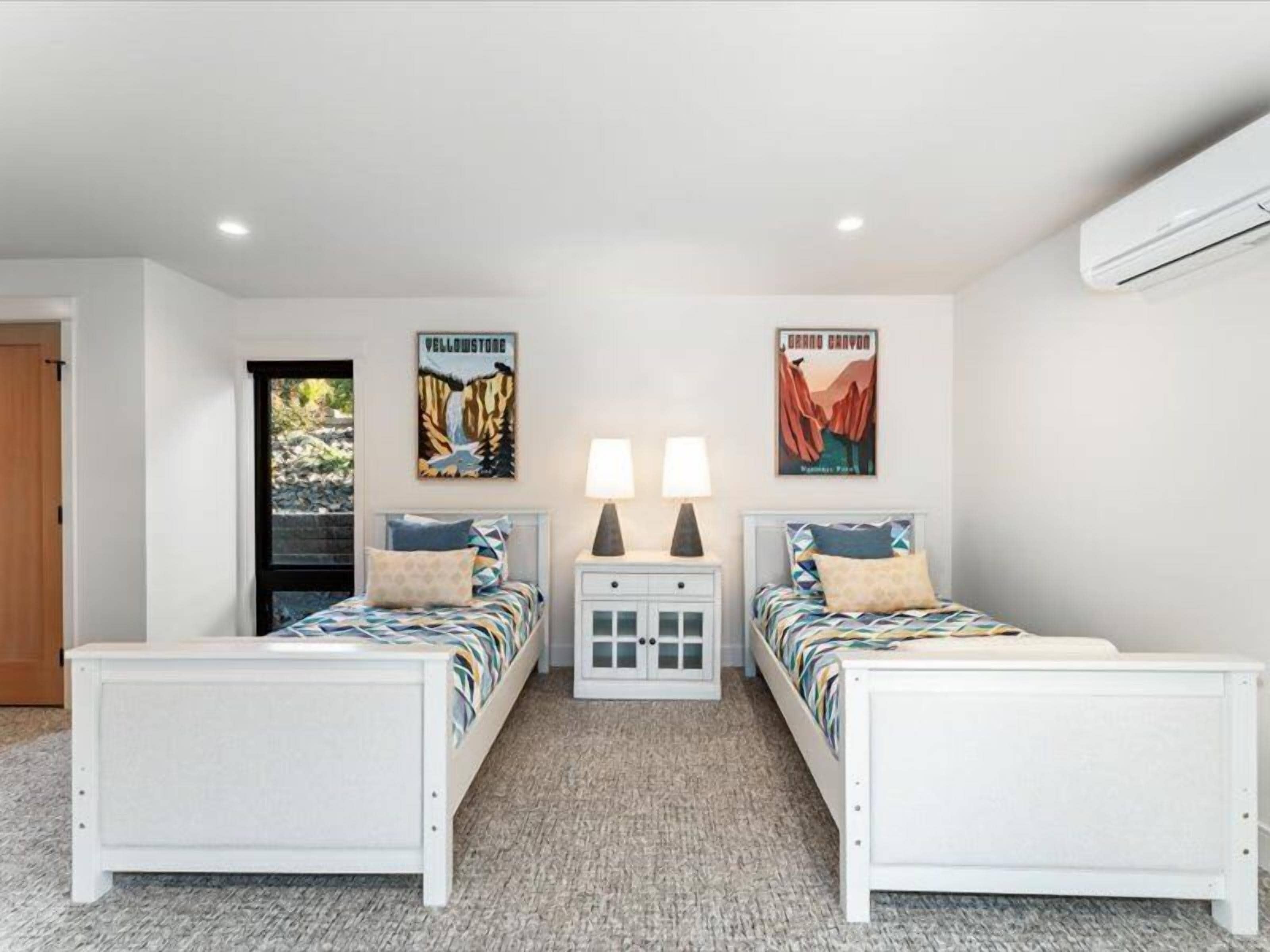 A bedroom with two beds, nightstands, wall art, and an air conditioning unit.