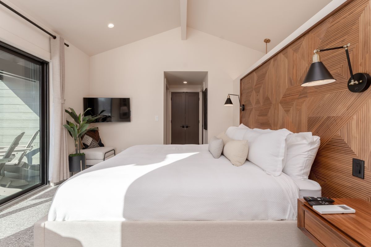 Een moderne slaapkamer met een groot bed, een houten hoofdbord, een nachtkastje, een aan de muur bevestigde televisie en uitzicht op het groen via een schuifdeur.