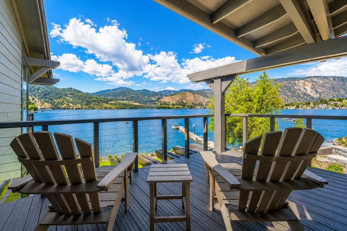 A deck with two Adirondack chairs and a table, overlooking a lake with mountains in the background.