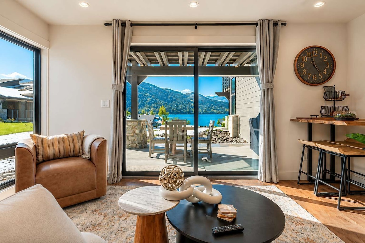 A modern living room with a leather sofa, a round coffee table with a decorative item, a dining area with a wooden table and chairs, and a large window with a mountain view.